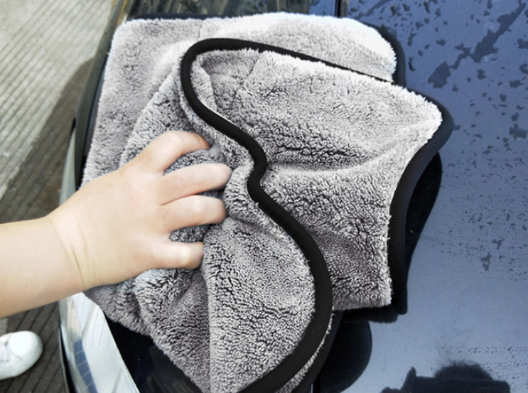 The 42 x 48 cm microfibre drying towel in dark grey with a black edge binding, held in one hand and wiped across a wet car panel.